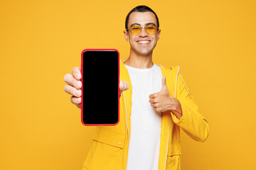 Young middle eastern man wear raincoat casual clothes sunglasses hold use mobile cell phone with blank screen workspace area show thumb up isolated on plain yellow orange background Lifestyle concept.
