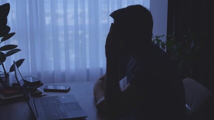 dark unrecognizable silhouette unhappy tired man working on laptop, sitting at desk. black room window light. Sad guy suffers from headache. depression fatigue lack of money. Person looking for job - Powered by Adobe