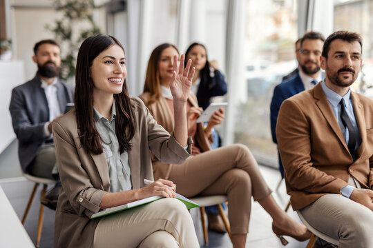 Smiling businesswoman sitting on seminar with her coworkers with hand raised and asking questions.