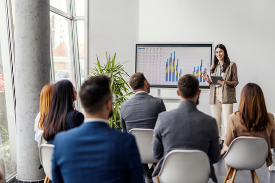 Young female data analyst is standing in boardroom in front of audience and presenting data statistics. - Powered by Adobe