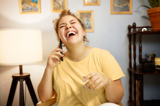 Cheerful puffy female with hair bun laughing out loud talking on phone with her bestie on her day off, gossiping and having fun to get positive mood, discussing epic fails at work or memes