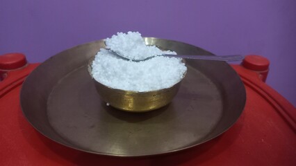 This image shows a metal bowl filled with white sago pearls (sabudana) placed inside a larger brass plate.A spoon rests inside the bowl, scooping some of the sago pearls.