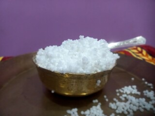 This image shows a metal bowl filled with white sago pearls (sabudana) placed inside a larger brass plate.A spoon rests inside the bowl, scooping some of the sago pearls.