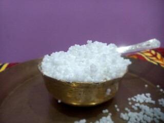 This image shows a metal bowl filled with white sago pearls (sabudana) placed inside a larger brass plate.A spoon rests inside the bowl, scooping some of the sago pearls.