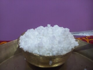 This image shows a metal bowl filled with white sago pearls (sabudana) placed inside a larger brass plate.A spoon rests inside the bowl, scooping some of the sago pearls.