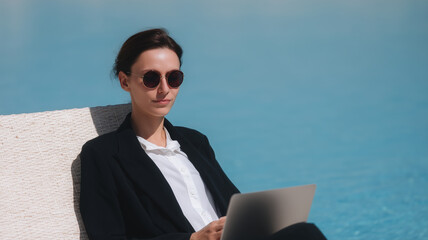 Relaxed professional woman in black suit and sunglasses working on laptop by luxurious blue poolside