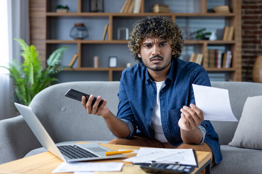 A concerned man sits in a living room holding a smartphone and bill, analyzing finances.