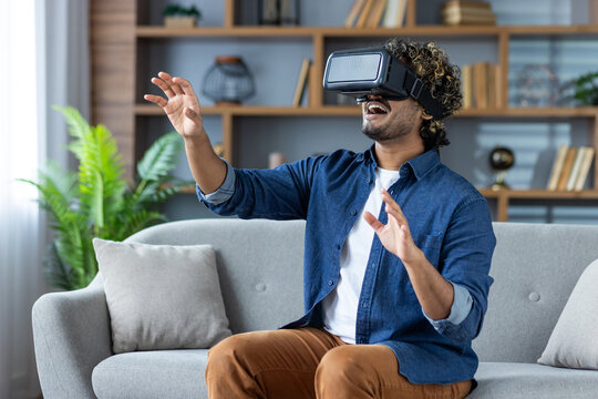 A man wearing a VR headset is exploring a virtual experience while seated on a sofa. The background features a neatly arranged bookshelf and indoor plants, creating a serene and modern atmosphere.
