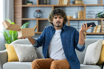 A man sitting on a couch, expressing confusion while holding a box and a smartphone. The setting is...