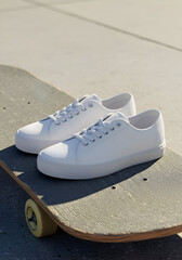 Pair of white canvas sneakers resting on a skateboard, outdoors. Casual streetwear style, youthful demographic, and urban setting.