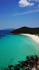 Fototapeta premium Serene coastal scene; turquoise water laps a white sand beach curving around a verdant hill under a clear blue sky