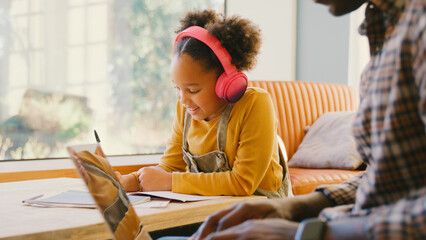 Father At Home Working On Laptop With Daughter Wearing Headphones As She Does Homework At Table