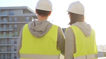 Two architects wearing safety hard hats and vests holding blueprint and discussing construction...