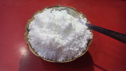 
A brass bowl filled with white sago pearls (sabudana) and a spoon rests on a red perforated surface, showing the pearls’ small, round texture clearly.
A brass bowl filled with white sago pearls (sa

