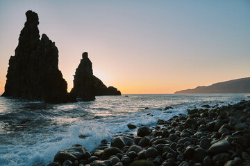 Majestic sea stacks at sunset on a rocky beach, with ocean waves crashing against the shore and distant coastal mountains under a warm, glowing sky.