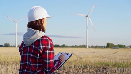 Female engineer wearing a white protective helmet is taking notes with a clipboard in a field with...