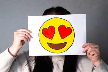 Woman holding heart eyed emoji mask in front of her face. Social media trends, Gen Z marketing, World emoji day 17 July.