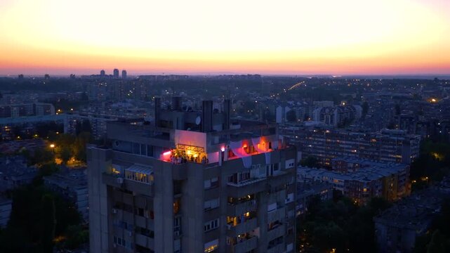 Aerial view of a rooftop party on a brutalist building in Belgrade at dusk. People dancing under red lights with the city skyline and Zemun quay in the background.