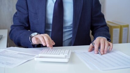 Businessman accountant with blue suit is using white calculator and taking notes with a silver pen....