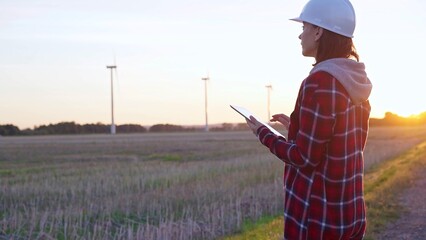 Woman engineer wearing a white protective helmet is taking notes with a tablet computer on a field...