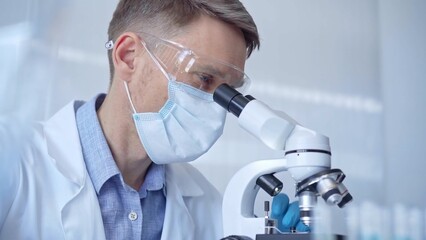 Man scientist with protective glasses, mask and gloves is using microscope in laboratory, portrait...