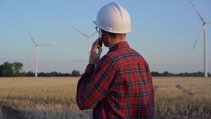 Man engineer, wearing a white protective helmet is talking by smartphone in a field with wind turbines, as the sun sets. Clean energy and engineering concept