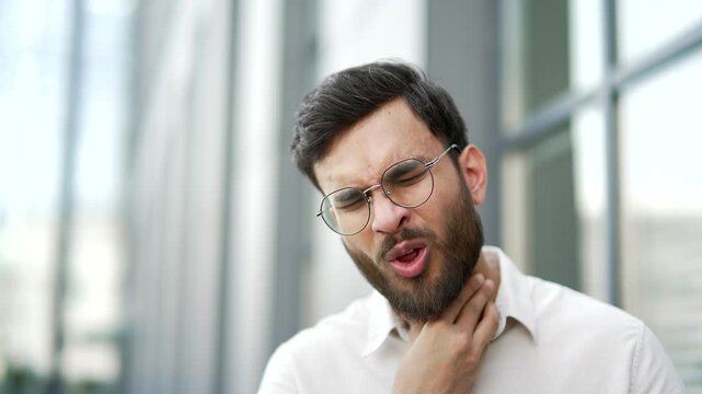 Sick businessman suffering from a sore throat while standing on street near business office building. Unhealthy man coughs, has a cold, virus or flu. Upset ailing male massaging a sore spot. Close up