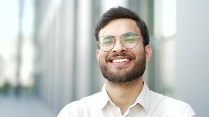 Portrait of smiling businessman in glasses standing on city street near office building. Happy bearded male entrepreneur looking at camera. Head shot of manager in shirt posing outdoors. Close up - Powered by Adobe