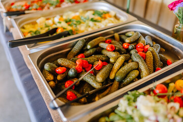 Close-up of a stainless steel buffet tray filled with pickles and red peppers, served with tongs at a catered event.