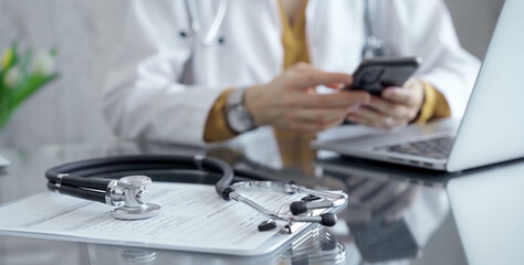 Close-up of doctor's desk with stethoscope and clipboard. Physician is using a tablet at the background. Medicine and health care