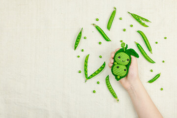 Hands holding crocheted pea toy among real pea pods on beige fabric. Natural colors