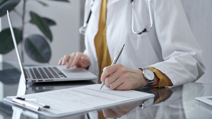 Doctor woman is writing on a document on a glass desk, with a laptop and tablet visible. Work space...