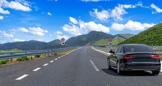 A sleek black car drives along a scenic mountain highway under a bright blue sky with scattered clouds