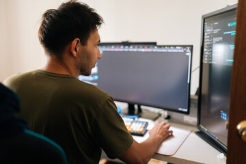 Close-up rear view of programmer male working from home office using multiple monitors setup and...
