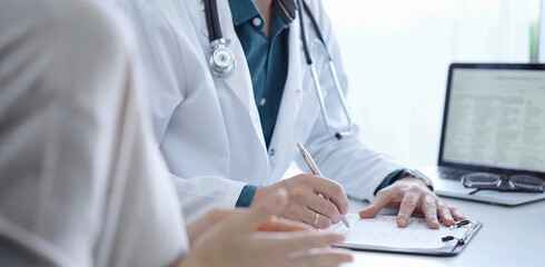 Doctor and a patient. The physician, wearing a white medical coat over a green shirt, is filling out a medical record form during a consultation in the clinic. Medical service