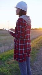 Woman engineer wearing a white protective helmet is taking notes with a clipboard in a field with...