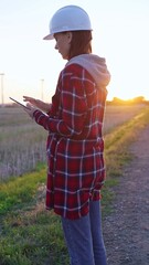 Woman engineer wearing a white protective helmet is taking notes with a clipboard in a field with...