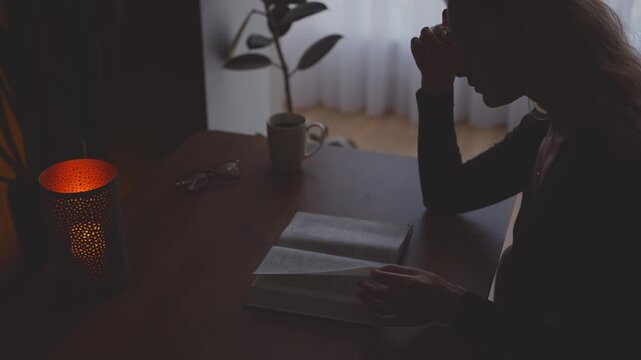 unrecognizable person dark silhouette woman reading book near window candle light black home room. girl sitting at desk holding paper book novel in hands, turning page. concept hobby rest relax 4k