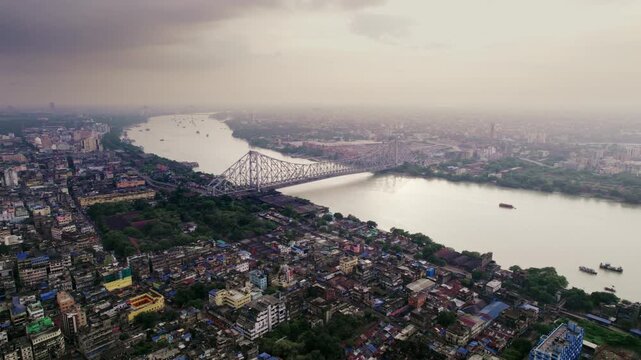 A cinematic daylight drone aerial shot of the historic heritage Howrah Bridge over the holy Hooghly or Ganges river, surrounded by Kolkata or Calcutta city with a vibrant cloudy sky in the background.