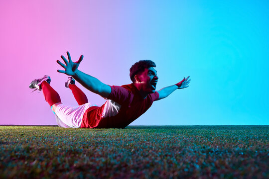 Soccer player sliding on grass celebrating victory in neon gradient lightning. Concept of high-energy sports joy, team triumph, and passionate success after winning goal.