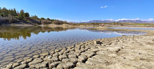 Low Water Level in a Cyprus Lake. A panoramic view of a dry lakebed in Cyprus with a low water level. Cracked mud fills the foreground, and mountains are visible on the horizon under a clear blue sky.