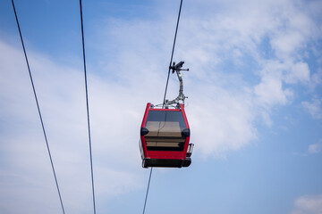 Red gondola cabin suspended on cableway in mountains. Alpine passenger transport. Cable car moving across blue sky over forest slopes
