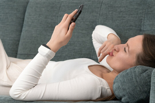 Young woman wearing a smartwatch is lying on a comfortable sofa at home, holding a smartphone above her and thinking with her finger on her lips