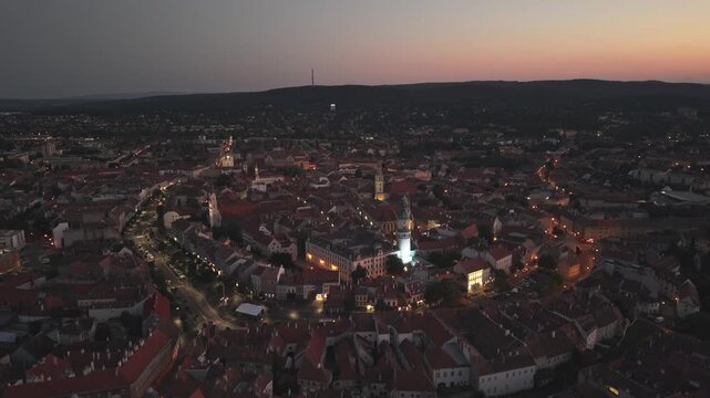 An aerial video of a historic European town during dusk, with warm streetlights illuminating the winding streets and iconic buildings. The fading light of sunset casts a soft glow over the rooftops.