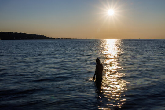 Silhouette of a person standing in calm sea waters during a golden sunset, with shimmering reflections and a tranquil horizon under a warm evening sky.