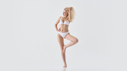 Fototapeta premium Blonde woman in white lingerie posing playfully with hand in hair and confident smile. Concept of fashion catalog, lingerie promotion, feminine charm, elegance and body positivity.