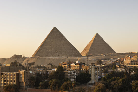 View of the majestic pyramids standing tall amidst the urban sprawl under a warm sky, contrasting ancient wonder with modern life, Cairo, Giza Governorate, Egypt.
