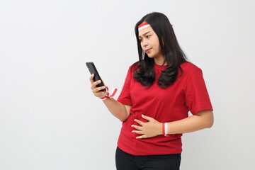 Naklejka premium Indonesian woman wearing red and white ribbon with confused expression choosing food using cellphone while holding her stomach, feeling hungry, ordering food online.