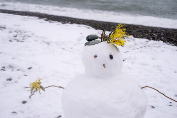 Cute snowman decorated with yellow mimosa flowers against the blue sea on a snowy beach in winter. Holiday background with cute snowman. Christmas card