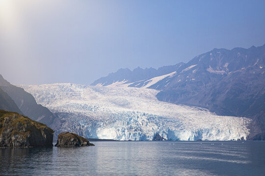 View of a massive blue glacier cascading towards the calm waters of Aialik Bay, nestled between rocky shores and snow-dusted mountains of Kenai Fjords National Park, Seward, Alaska, United States.